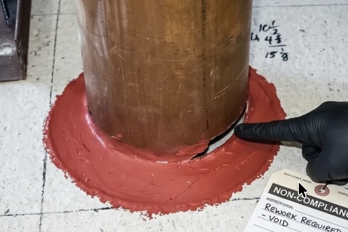 Inspector pointing to a crack in red firestop sealant around a copper pipe penetration through a floor, with a non-compliance rework required tag visible