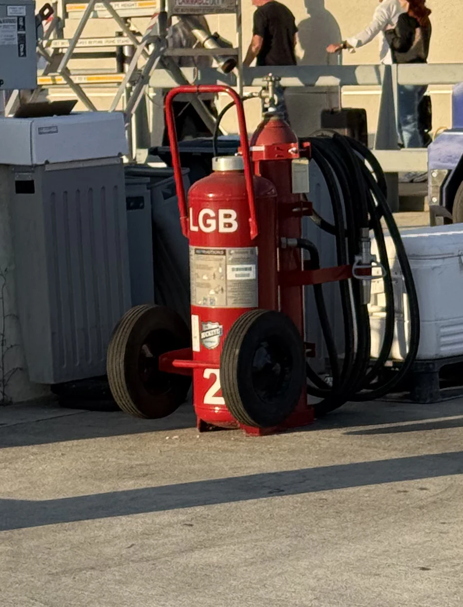 A red wheeled cartridge-operated dry-chemical fire extinguisher with two large spoked wheels, an external nitrogen expellant cartridge, and a coiled discharge hose, stenciled LGB, staged beside fuel-servicing cabinets at an airport gate.